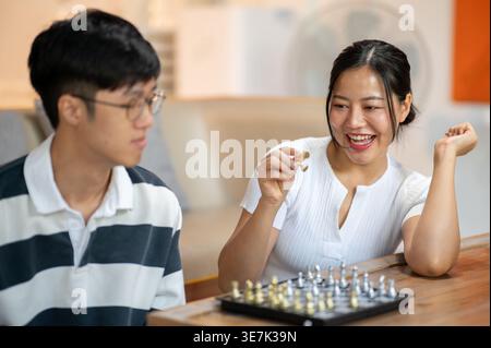 Asiatische Frau in weißem Hemd, die Schachstücke hält, während sie mit einem männlichen Freund im Wohnzimmer zu Hause Schach spielt Stockfoto