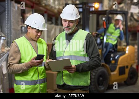 Gemischte weibliche und kaukasische Arbeiter halten Klemmbrett und reden, während sie im Lager stehen Stockfoto