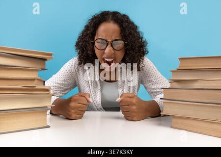 Gestresste schwarze Teenagerin mit Brille und schreiender Brille, während sie im Studio mit Bücherstapeln vor blauem Hintergrund am Tisch sitzt Stockfoto