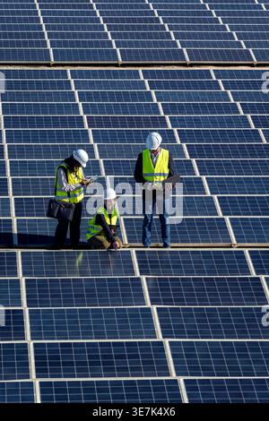 Verschiedene Ingenieure, die Schutzhelme und Sicherheitswesten mit Klemmbrett, Laptop und Tablet tragen, während sie große Solarpaneele beim Bau inspizieren... Stockfoto