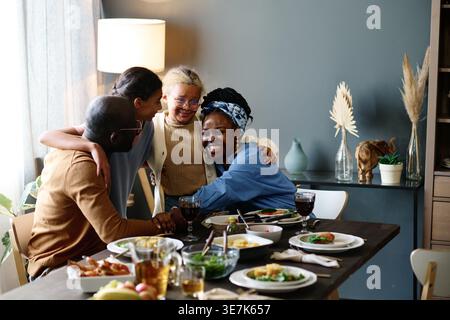 Schwarze Eltern haben Spaß, ihre Töchter zu umarmen, während sie zu Hause bei Essen und Getränken am Tisch sitzen Stockfoto