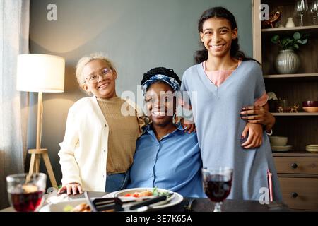 Schwarze Mutter trägt ein Stirnband, das ihre Töchter umschließt, während sie zu Hause beim Essen und Trinken am Tisch sitzt Stockfoto