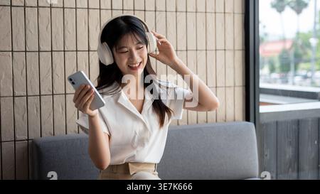 Thailändische Frau, die ihr Smartphone hält und Musik über Kopfhörer hört, während sie im Café auf der Couch sitzt Stockfoto