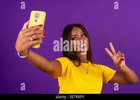 Hispanische Frau zeigt Friedenszeichen und macht Selfie mit Smartphone, während sie im Studio vor violettem Hintergrund steht Stockfoto