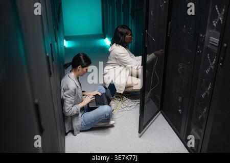 Schwarze IT-Ingenieurin mit Brille in der Nähe eines kaukasischen Kollegen, der Laptop benutzt, während sie Kabel im Serverschrank hockt und verbindet... Stockfoto