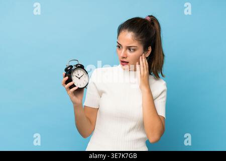 Hispanische Frau mit schockiertem Ausdruck, während sie auf den Wecker vor blauem Hintergrund in stduio blickt Stockfoto