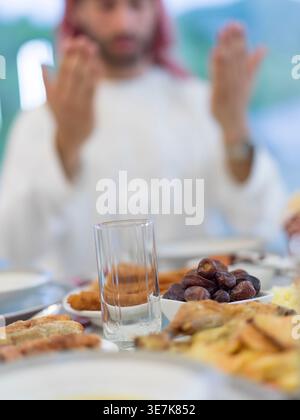 Mann aus dem Nahen Osten in traditioneller arabischer Kleidung betet während des Ramadan am Tisch mit Speisen im Speisesaal zu Hause Stockfoto