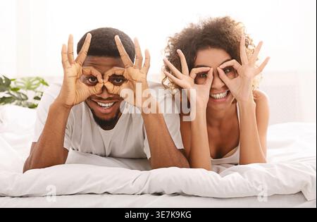 Fröhliches schwarzes Paar macht Handbrillen, während es zu Hause auf dem Bett liegt Stockfoto