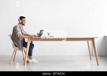 Fröhlicher Mann aus dem Nahen Osten, der Kopfhörer trägt, während er am Laptop arbeitet und am Tisch im Büro sitzt Stockfoto