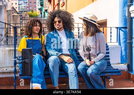 Hispanischer Mann mit Afro, der Jeansjacke und Sonnenbrille trägt und sich mit seinen Freundinnen unterhalten hat, während er zusammen auf einer blauen Bank in der Stadt sitzt... Stockfoto
