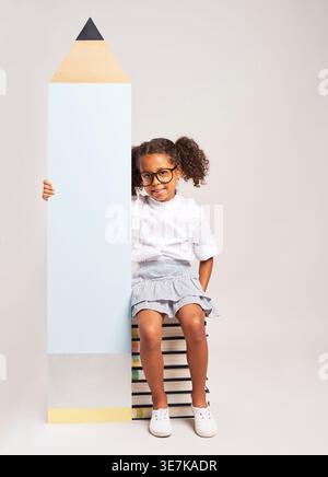 Schwarzes Mädchen mit lockigem Haar, Brille, die auf einem Stapel Bücher sitzt und großen Bleistift vor grauem Hintergrund im Studio hält Stockfoto