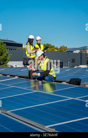 Verschiedene Ingenieure, die Schutzhelme und Sicherheitswesten mit Klemmbrett, Laptop und Tablet tragen, während sie große Solarpaneele beim Bau inspizieren... Stockfoto