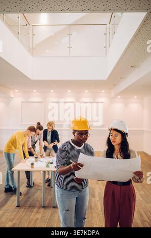 Serious Black Woman und asiatische Frauen mit Schutzhelmen, die im Büro an einem Entwurf arbeiten Stockfoto