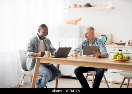 Schwarzer Mann, der den Becher hält und ein Tablet benutzt, während er am Tisch sitzt, mit einem männlichen Freund mittleren Alters, der in der Küche zu Hause am Laptop arbeitet Stockfoto