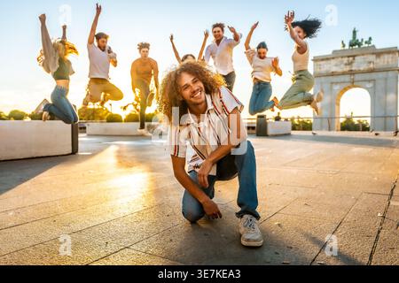 Lächelnder hispanischer Mann mit lockigen Haaren, der sich während des Sonnenuntergangs vor verschiedenen Freunden in der Stadtstraße hockt Stockfoto