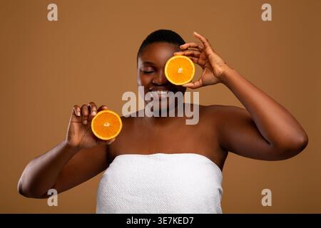 Schwarze Frau im Badetuch bedeckt Auge mit Zitrusfrüchten, die vor braunem Hintergrund im Studio stehen Stockfoto