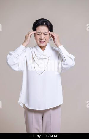 Gestresste chinesische Seniorin mit Kopfschmerzen, die im Studio mit Händen auf der Stirn auf braunem Hintergrund steht Stockfoto