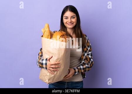 Hispanische Frau mit Papiertüte voller Brot, die vor violettem Hintergrund im Atelier steht Stockfoto