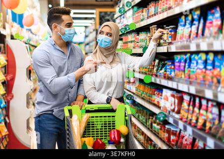 Ein lächelndes Paar aus dem Nahen Osten, das Essen aus den Regalen wählt, während er mit dem Wagen im Supermarkt steht Stockfoto