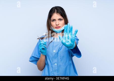 Hispanische Zahnärztin, die Zahnspiegel und Sichelsonde hält, während sie vor hellblauem Hintergrund steht Stockfoto