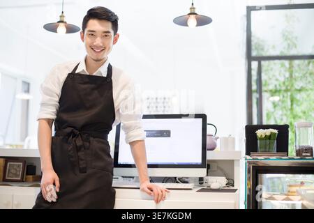 Chinesischer Mann mit Schürze, der am Tresen mit Computer im Café steht Stockfoto