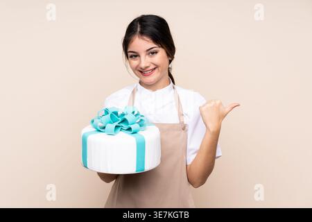 Hispanische Konditorin, die den Daumen zur Seite zeigt und im Studio den Kuchen vor beigem Hintergrund hält Stockfoto