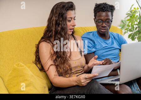 Schwarze männliche und kaukasische Paare arbeiten gemeinsam an ihren Haushaltsfinanzen und prüfen Papierdokumente, während ein Laptop zu Hause auf der Couch sitzt Stockfoto