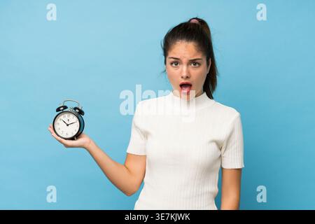 Hispanische Frau mit schockiertem Ausdruck, während sie den Wecker vor blauem Hintergrund in stduio hält Stockfoto