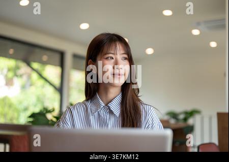 Thailändische Frau, die wegschaut und am Tisch mit Laptop im Café sitzt Stockfoto