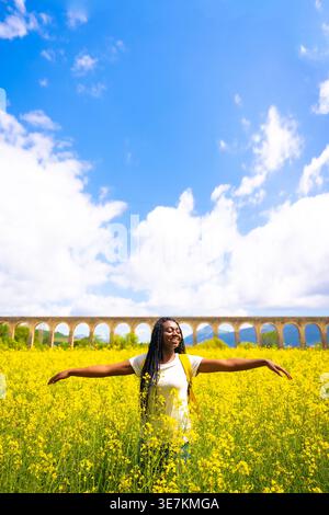 Lächelnde schwarze Frau mit Wendungen, die mit ausgestreckten Armen auf einem Feld mit gelben Blumen an sonnigem Tag steht Stockfoto