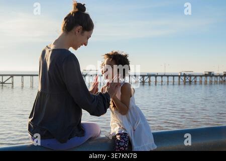 Hispanische Mutter und Tochter haben Spaß beim Spielen auf dem Straßenzaun in der Nähe des Piers bei Sonnenuntergang Stockfoto
