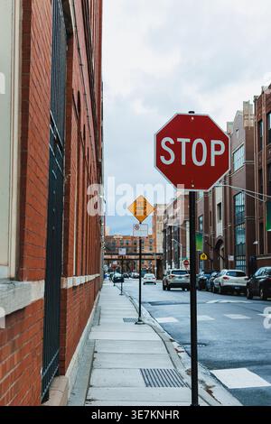 Ein Stoppschild steht am Rande eines Bürgersteigs neben einem Backsteingebäude. Ein Schild mit Geschwindigkeitsbegrenzung ist weiter unten auf der Straße zu sehen. Autos parken entlang der Straße Stockfoto