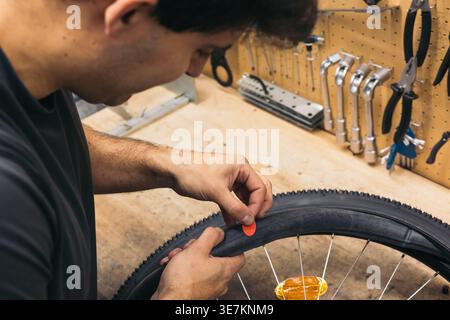 Hispanischer männlicher Mechaniker, der während des Aufstehens in der Werkstatt einen roten Fleck auf einen durchbrochenen Fahrradreifen legt Stockfoto