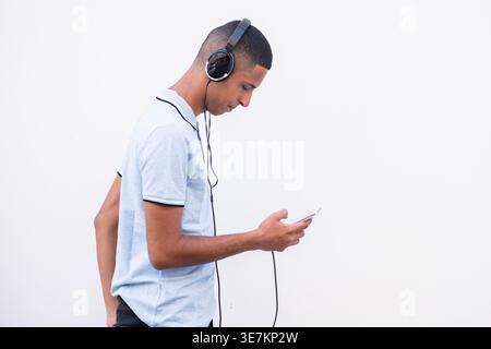 Arabischer männlicher Teenager in Kopfhörern mit Smartphone stehend mit Hand in der Gesäßtasche vor weißem Hintergrund Stockfoto