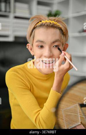 Lächelnder asiatischer schwuler Mann Aufnahme Make Up Tutorial Anwendung Lidschatten sitzend auf dem Boden in der Nähe des Sofas zu Hause Stockfoto