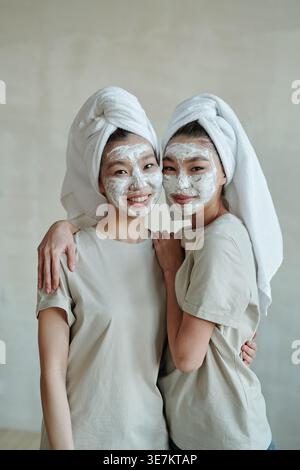 Asiatische Schwestern mit Gesichtsmasken und Köpfen, die in Handtücher gehüllt sind und sich zu Hause an die graue Wand schmiegen Stockfoto