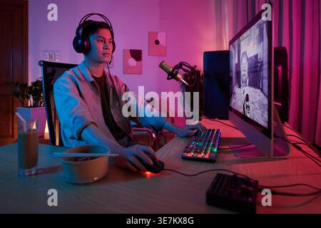 Asiatischer männlicher Online-Game-Streamer, der ein Headset trägt, das auf dem Computer spielt, während er zu Hause auf einem Stuhl sitzt Stockfoto