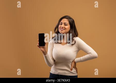 Glückliche junge arabische Frau, die modernes Smartphone mit leerem Bildschirm zeigt, eine mobile App empfiehlt oder ein fantastisches Angebot auf braunem Studiohintergrund zeigt, moc Stockfoto
