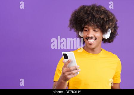 Lächelnder Mixed Race man mit Afro, der Musik mit Kopfhörern anhört, und das Smartphone steht vor violettem Hintergrund Stockfoto