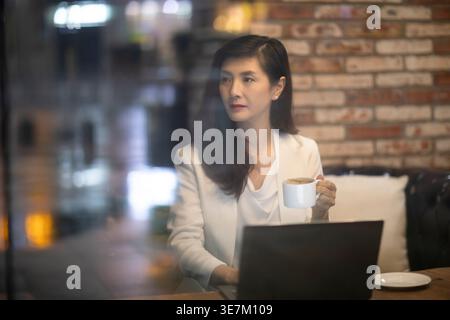 Chinesische Frau, die eine Tasse Kaffee hält und wegschaut, während sie am Tisch im Café sitzt Stockfoto