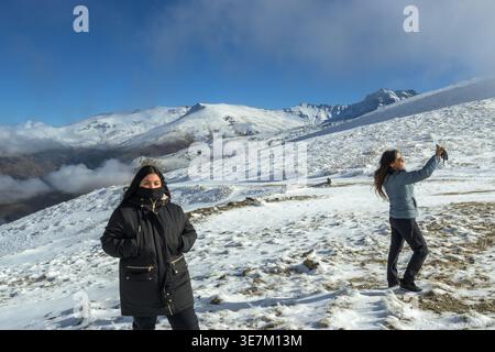Hispanische Frau in warmer Jacke, die neben ihrer Mutter steht und im Winter Fotos auf schneebedecktem Feld macht Stockfoto