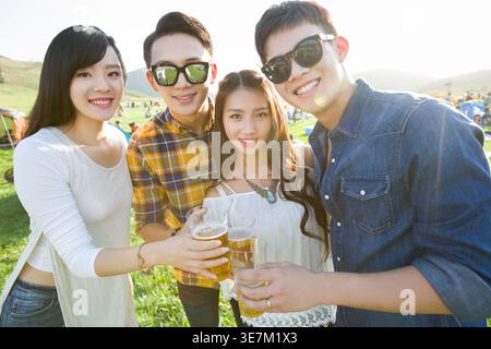 Lächelnde chinesische Freundinnen und Freundinnen, die beim Musikfestival zusammen auf dem Konzertgelände mit Zelten eine Gläser mit Bier anschlagen Stockfoto