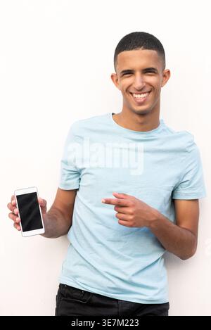 Lächelnder arabischer männlicher Teenager, der mit dem Finger auf das Smartphone zeigt, das vor weißem Hintergrund steht Stockfoto