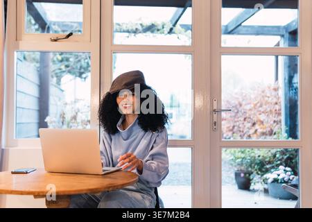 Lächelnde Mixed Race Frau mit lockigem Haar, die Baskenmütze mit Laptop trägt, während sie zu Hause am Tisch vor der Glaswand sitzt Stockfoto