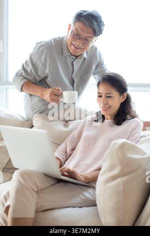 Lächelndes chinesisches Paar mittleren Alters, das zu Hause auf dem Sofa sitzt Stockfoto