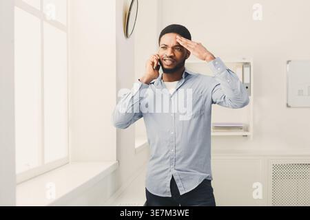 Verwirrter Schwarzer, der telefoniert, seine Stirn berührt und nachdenklich wegblickt, während er zu Hause steht Stockfoto