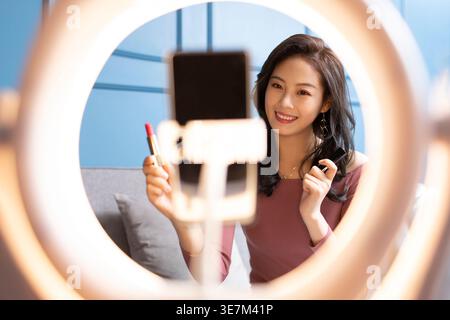 Lächelnde Chinesin, die Lippenstift hält, während sie zu Hause Live-Streaming macht Stockfoto