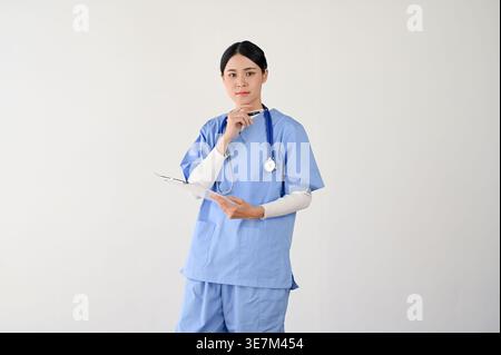 Asiatische Ärztinnen in Peelings, die Stethoskop um den Hals tragen und das Kinn berühren, das Clipboard vor grauem Hintergrund hält Stockfoto