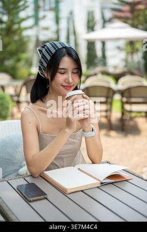 Eine attraktive asiatische Frau in einem süßen Kleid riecht ihren Kaffee, die Augen geschlossen, genießt ihren Kaffee, während sie an einem hellen Tag in einem wunderschönen Garten entspannt. Stockfoto