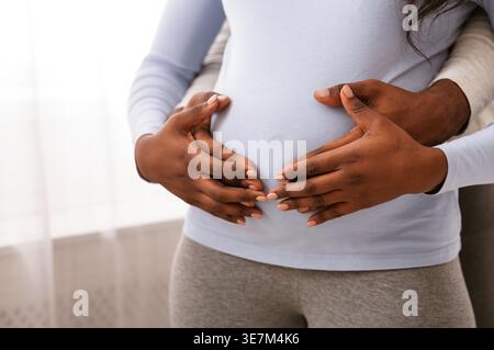 Abgeschnittenes Bild eines Schwarzen Mannes, der hinter einer schwangeren Frau steht, während er tagsüber den Bauch im Schlafzimmer berührt Stockfoto